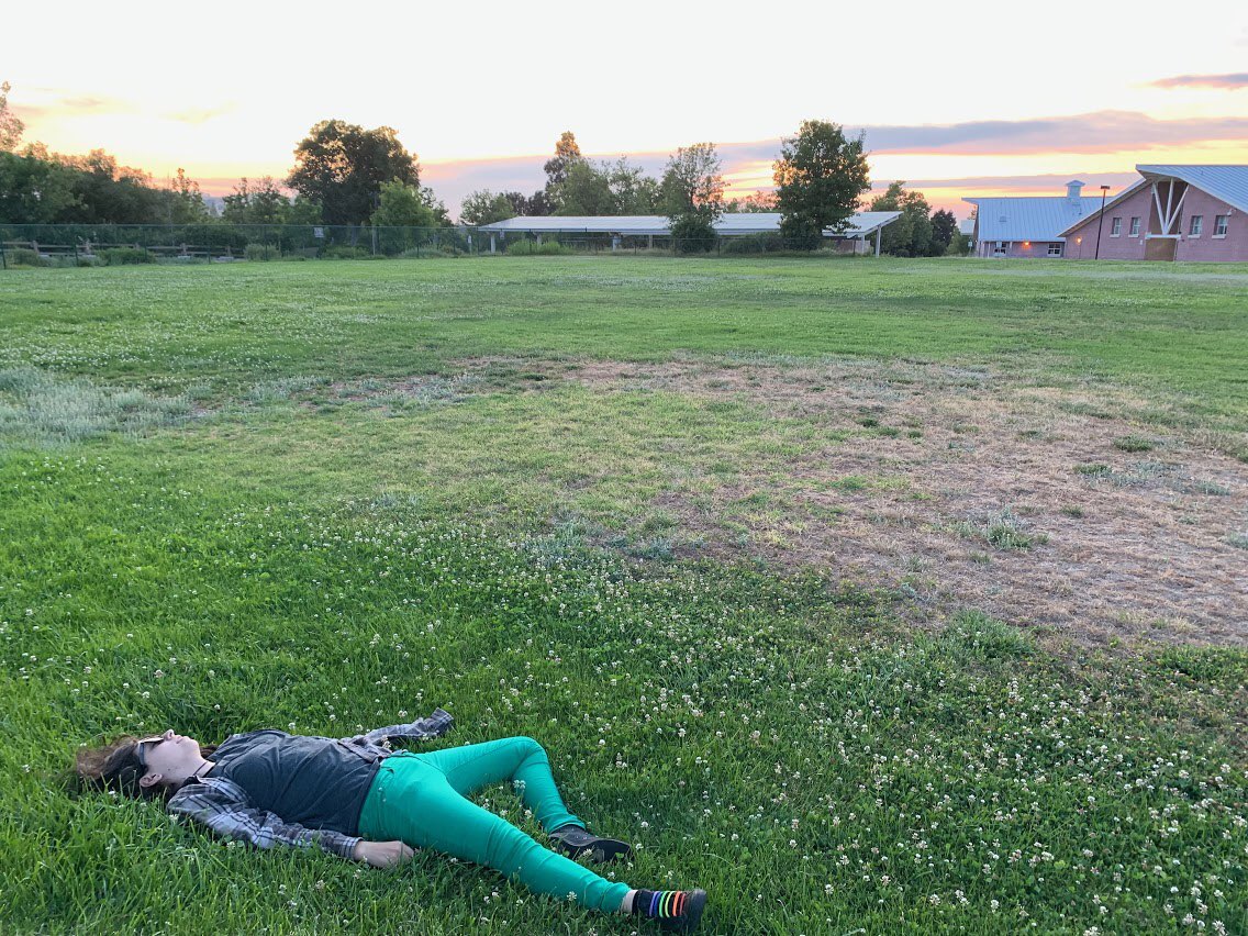 Image ID: A photo of a person lying in a grassy field, overlooking a sunset, with buildings visible in the background. They are wearing sunglasses, green pants, and shoes with rainbow laces. Their face is turned so it is barely visible.